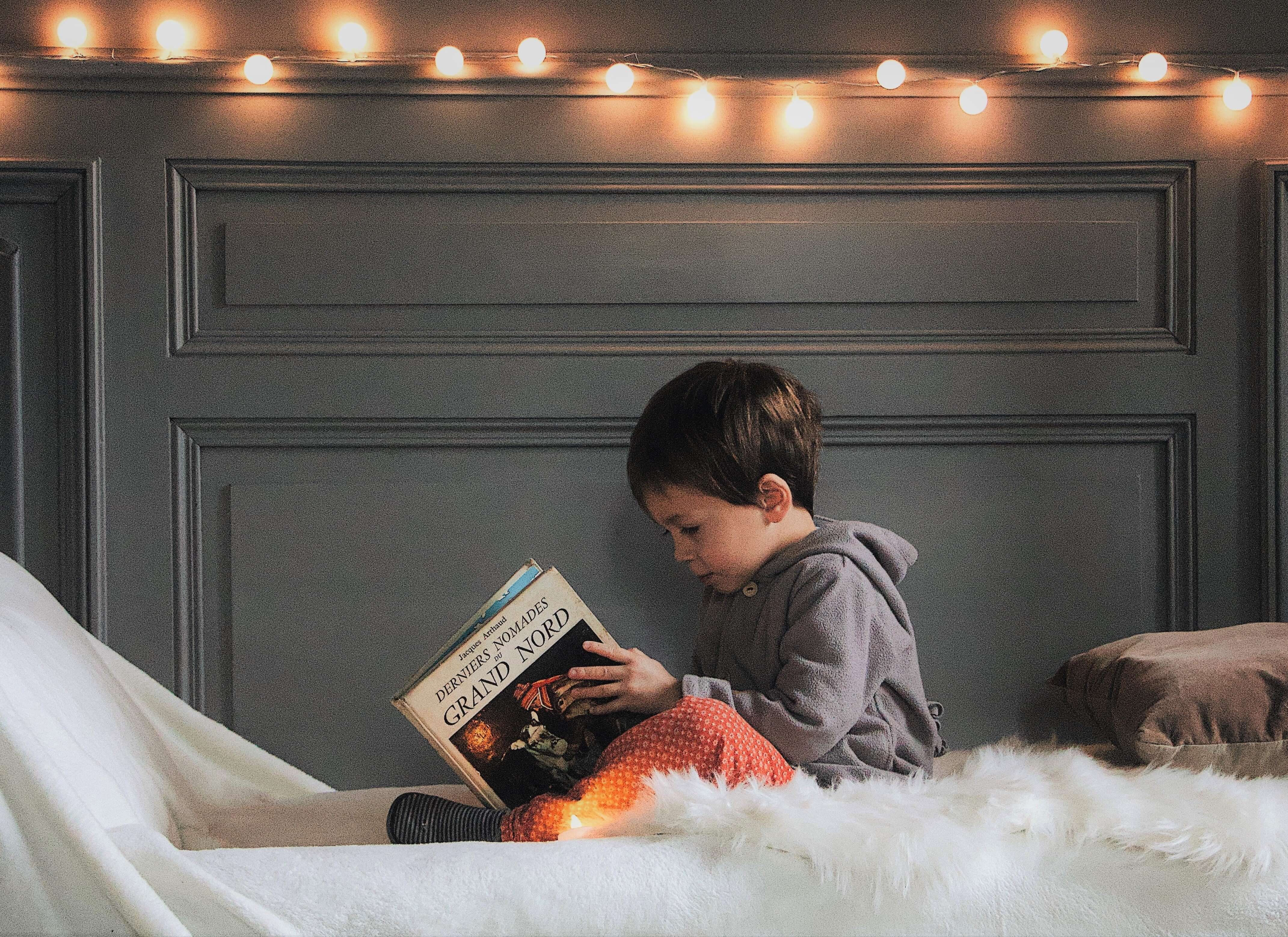 Jongetje leest boek op bed, lichtjesslinger boven zijn hoofd aan de grijze gelambriseerde wand