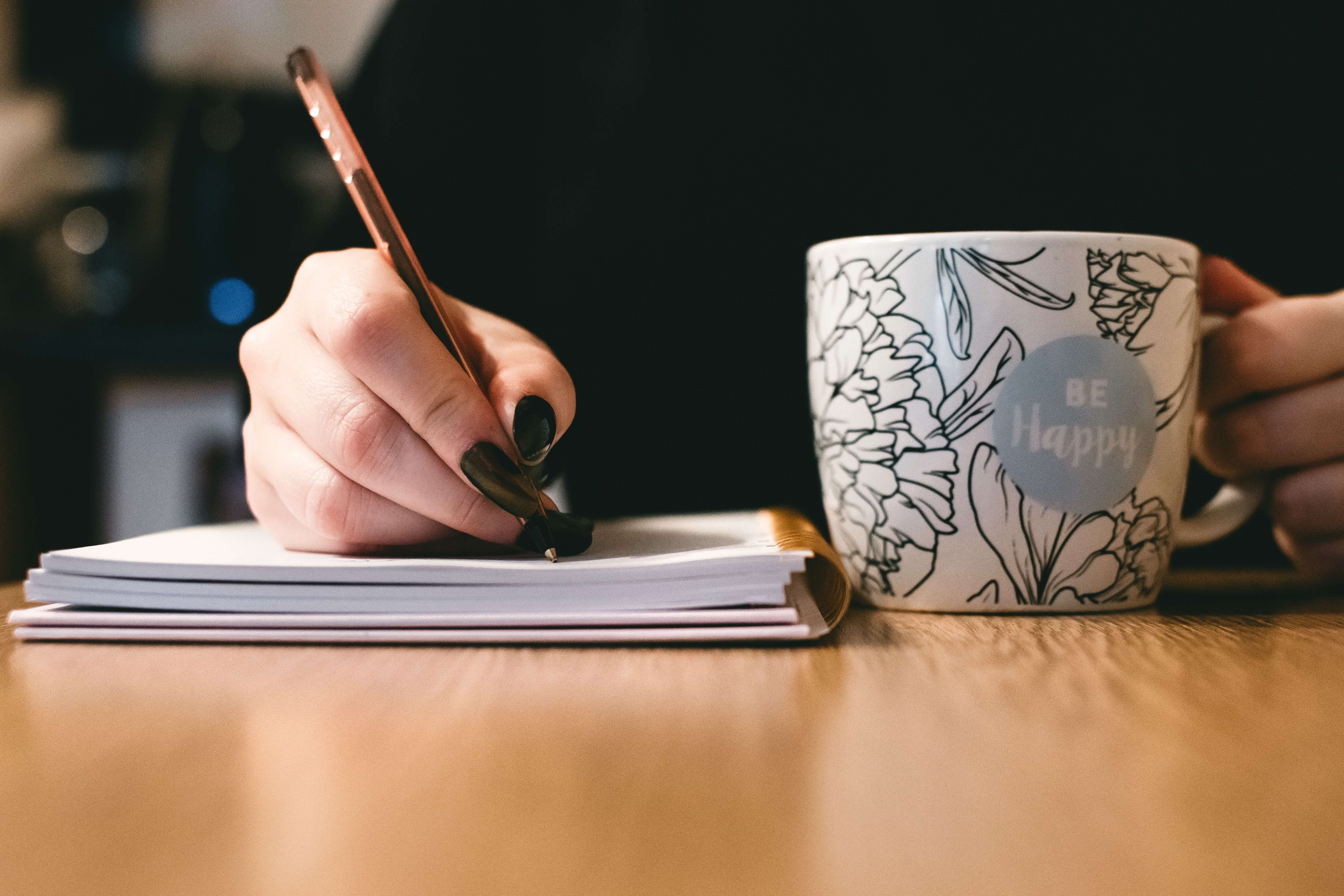 Vrouw schrijft met pen en papier en drinkt thee uit mok met tekst be happy