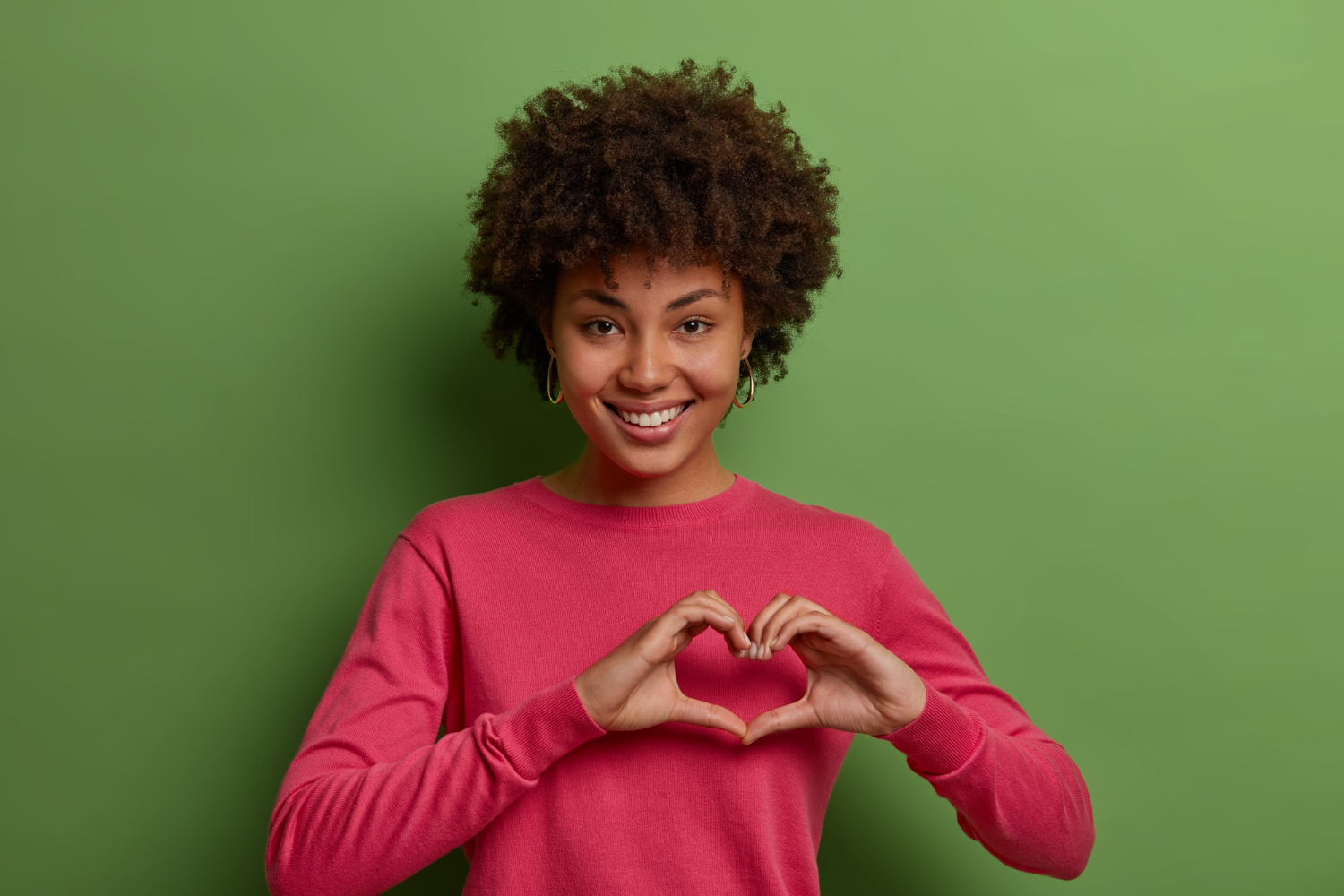 Zwarte vrouw met afrokapsel en roze trui met lange mouwen, vormt met haar handen een hart