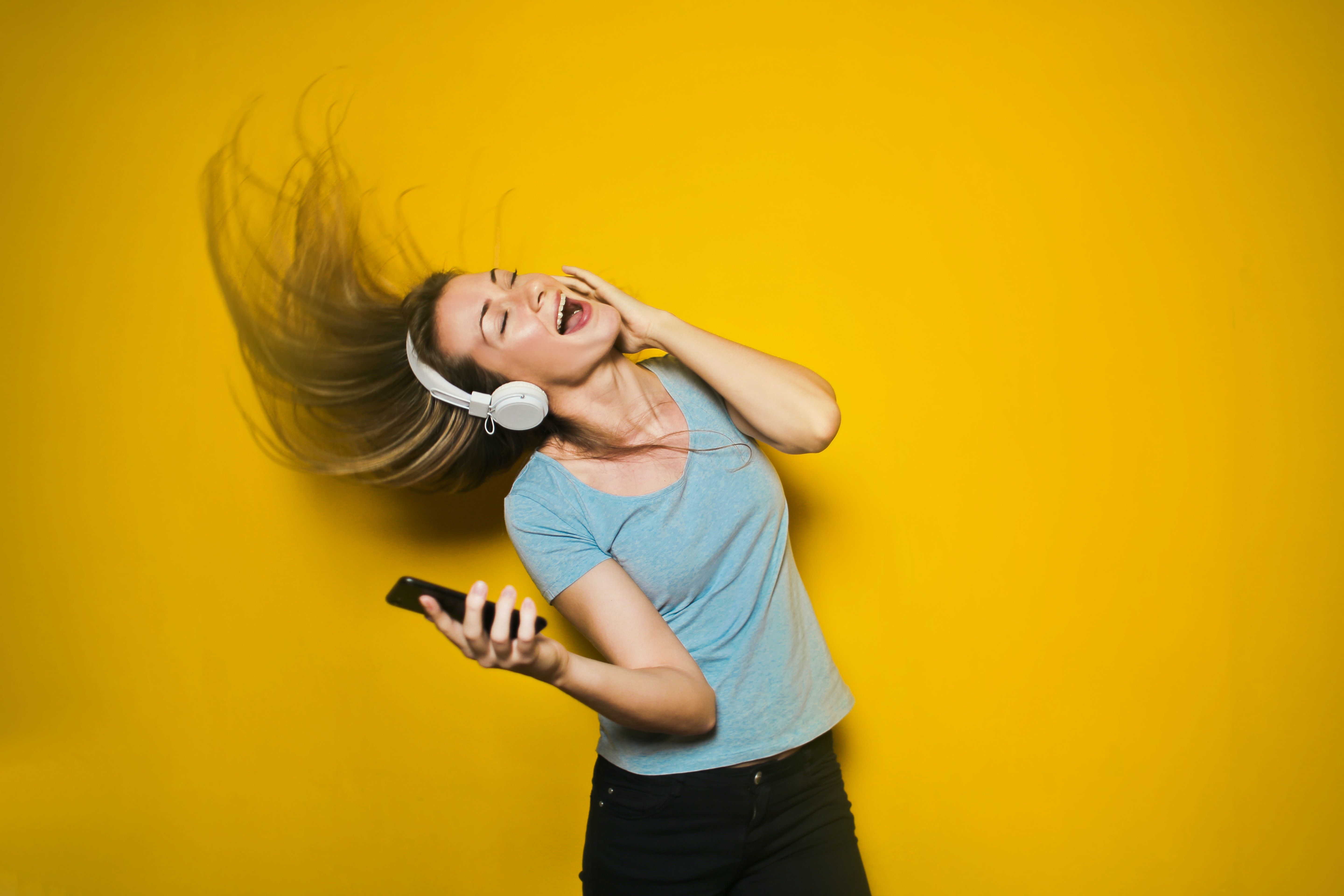 Vrouw in blauwe t-shirt met koptelefoon danst, gele achtergrond